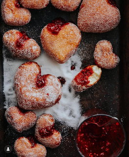 Heart-shaped jam doughnuts with powdered sugar and a bowl of red jam on a dark surface.