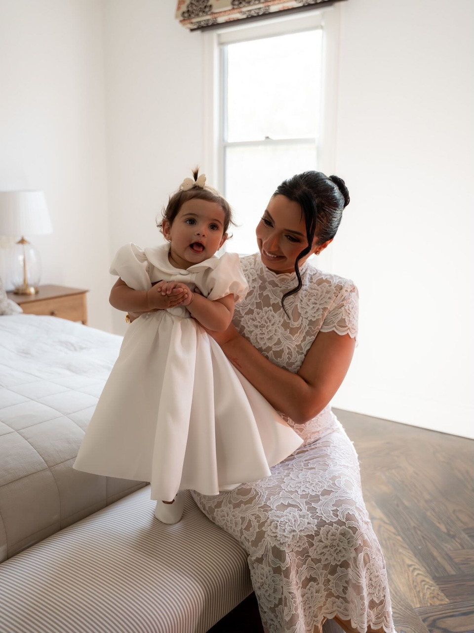 Woman in a white lace dress holding a baby in a white christening gown in a bedroom