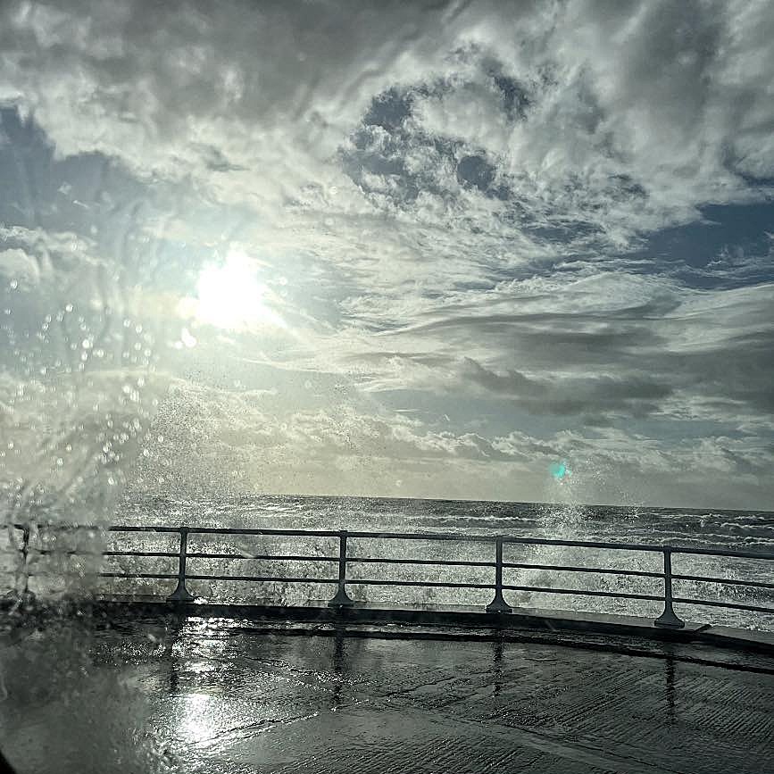 Large waves crashing over a metal railing with dramatic cloudy sky and sunlight breaking through