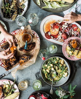 Overhead flatlay of abundant brunch spread with colorful plates of food, drinks, and hands reaching for dishes