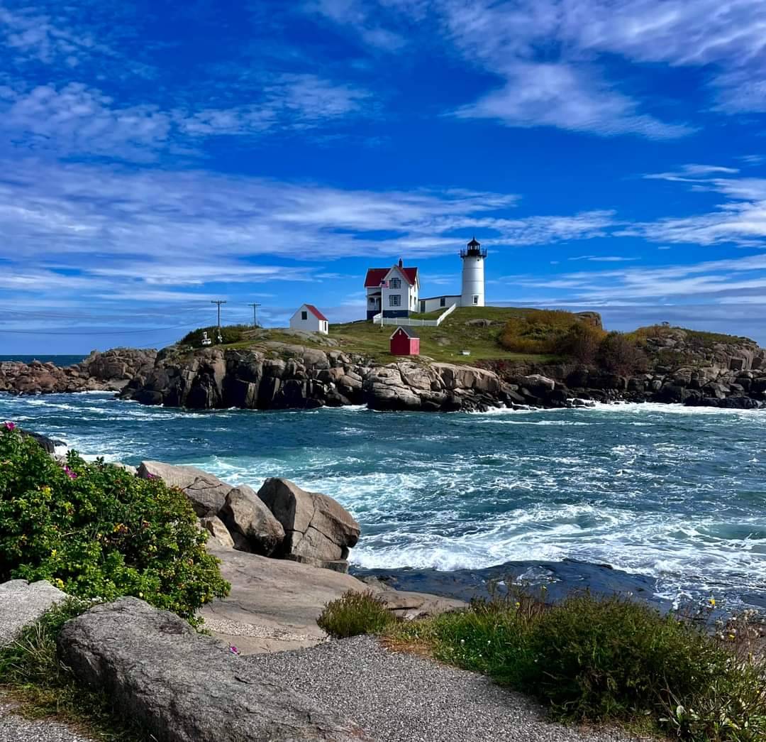 White lighthouse on rocky island with buildings, surrounded by ocean waves and blue sky