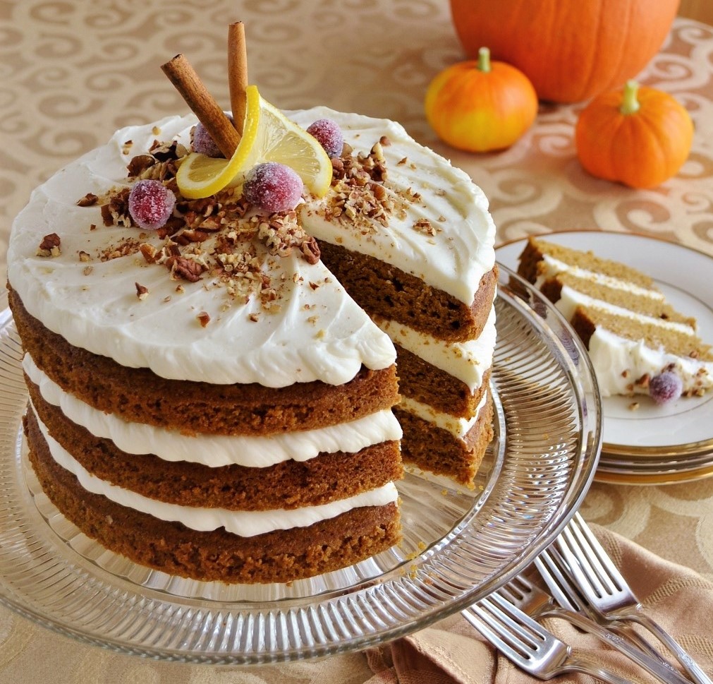 Layered pumpkin spice cake with white frosting, dried fruit, cinnamon sticks, and decorative pumpkins on a glass plate