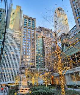 New York City skyscrapers surrounding a plaza with illuminated trees under a blue sky