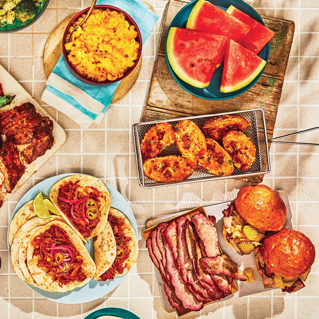 Overhead view of summer meal with grilled food, watermelon, corn, and burgers on checkered tablecloth