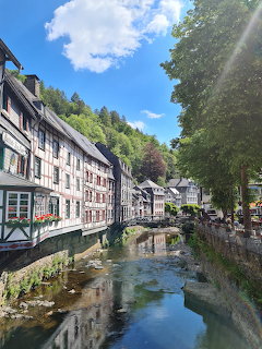 Half-timbered houses along a river in Monschau, Germany with green forested hillside and blue sky