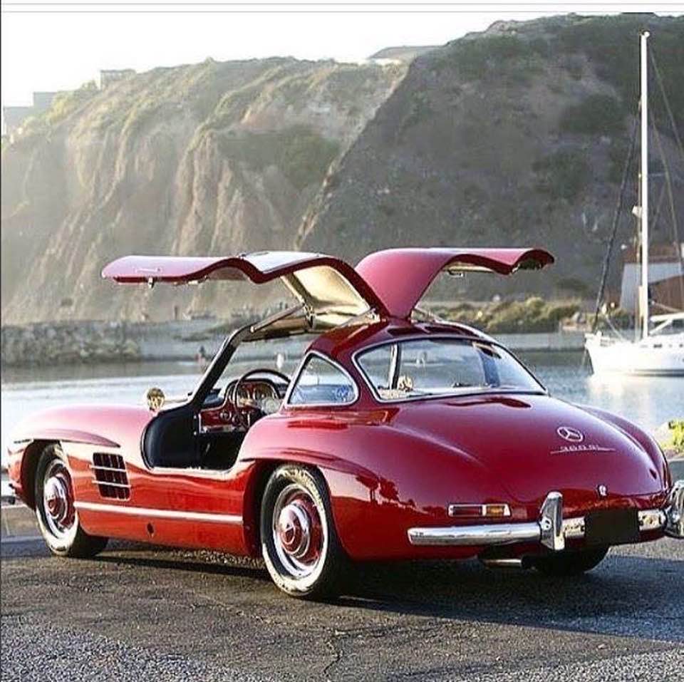 Red Mercedes 300SL Gullwing with open doors parked at waterfront with cliffs behind
