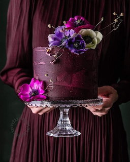 Person holding a deep purple cake topped with purple and cream flowers on a glass stand