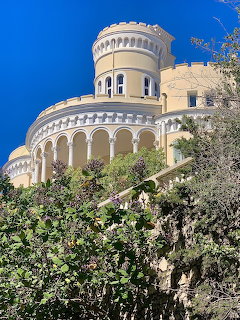 Grand cream-colored villa with round tower and arched columns surrounded by green trees under blue sky