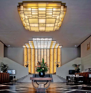 Art deco hotel lobby with illuminated glass ceiling, twin staircases, and checkered marble floor