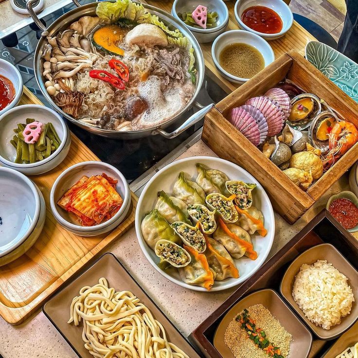 Overhead view of a Korean meal with hot pot, dumplings, kimchi, seafood steamer, noodles, and side dishes