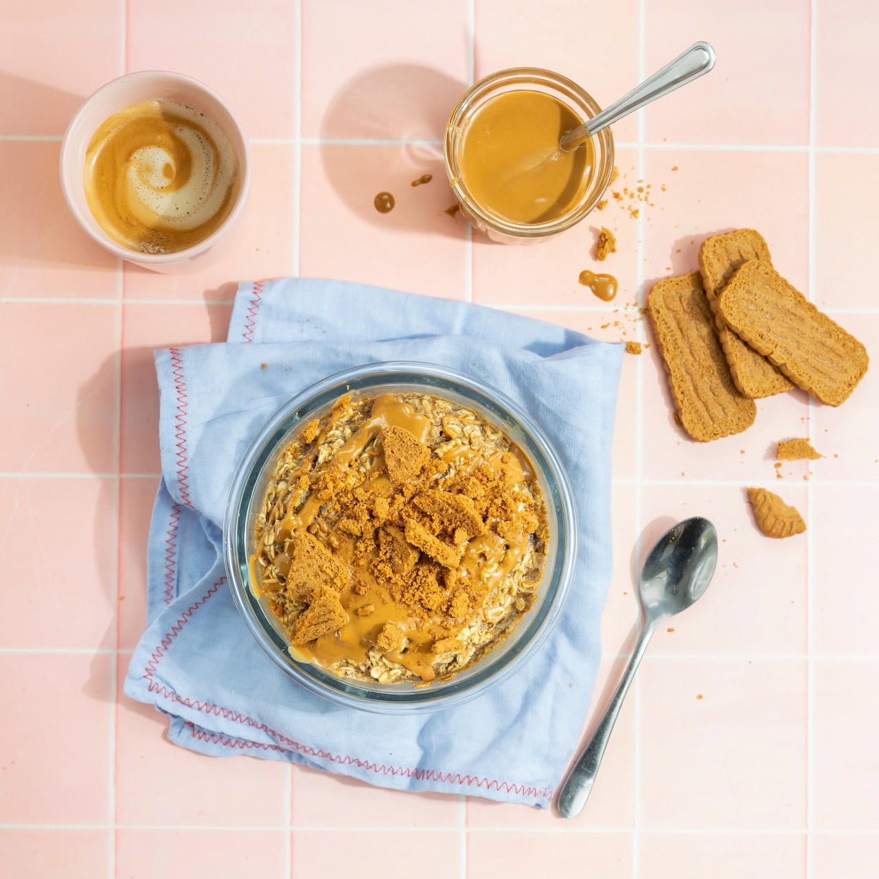 Top-down view of a bowl of biscuit crumble dessert with tea, cookies, and a spoon on pink tiles.