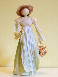 Handmade corn husk doll wearing a green pleated dress, white apron, and straw hat, holding a flower basket