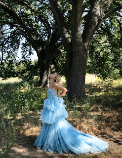Woman in flowing blue tiered gown standing by oak tree in sunlit meadow