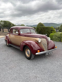 Vintage 1938 Hudson Big Six Coupe in maroon and tan parked on a driveway with rolling green hills behind