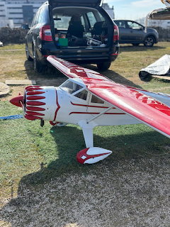 Red and white RC model airplane on grass with parked cars in the background