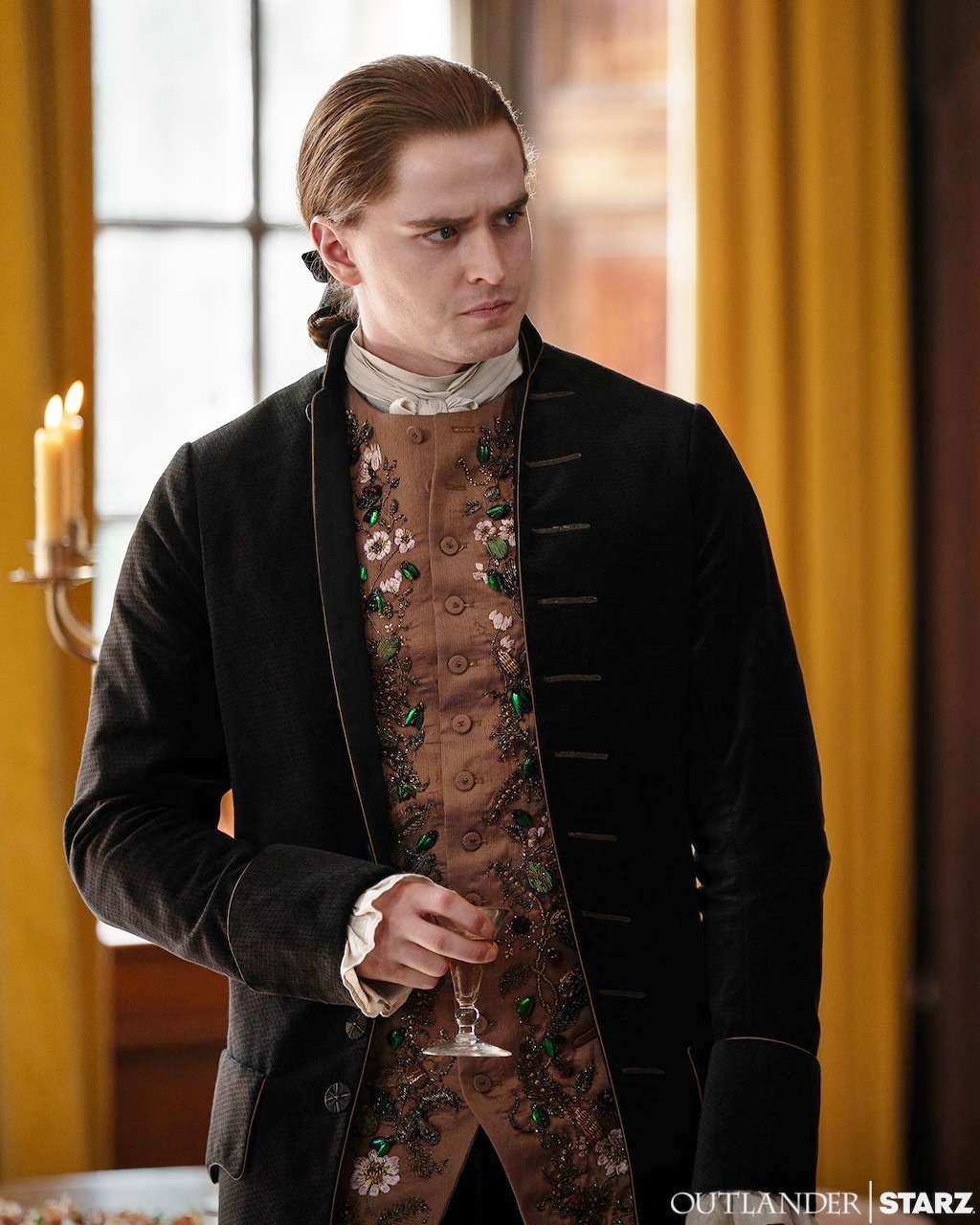 Man in 18th century black velvet jacket and brocade vest standing indoors in period drama costume