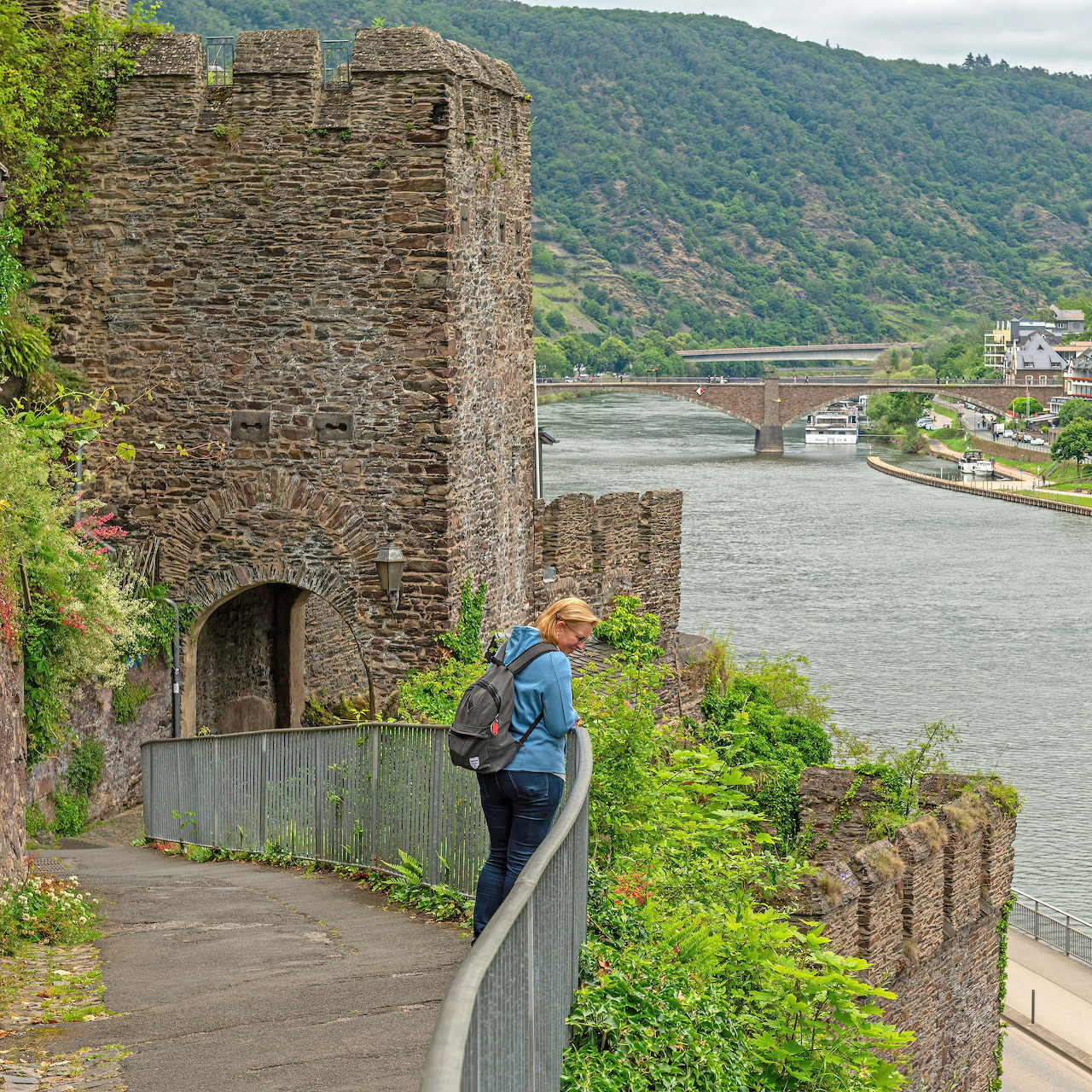 Person on walkway by stone tower ruins overlooking Moselle River and green hills at Cochem, Germany
