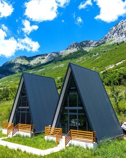 Two A-frame cabins on green hillside with mountains behind in Theth, Albania