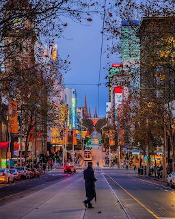 Person walking down Bourke Street in Melbourne at dusk with lit shops and tram tracks