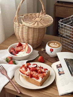 Strawberry toast, sliced strawberries, and a latte on a wooden table beside baskets and an open magazine.