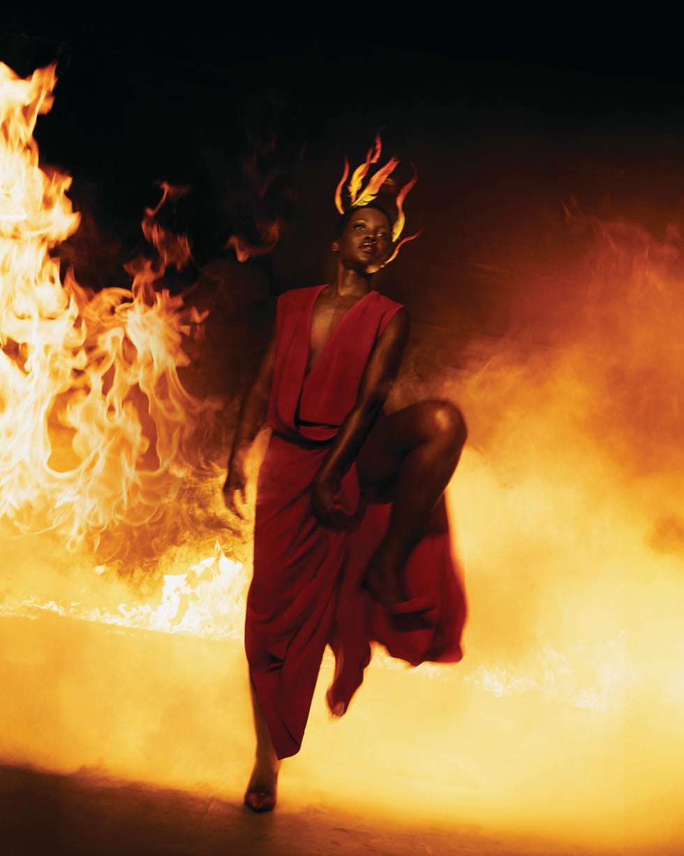Woman in red flowing dress striding through large flames and orange smoke in a dramatic fashion shoot