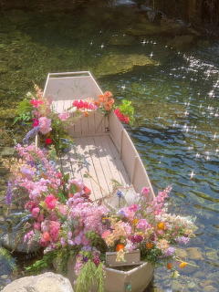 Small white wooden boat overflowing with pink, purple, and orange flowers floating on a sparkling pond