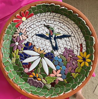 Circular mosaic birdbath with blue bird center, white daisy, and colorful flower tiles around the rim