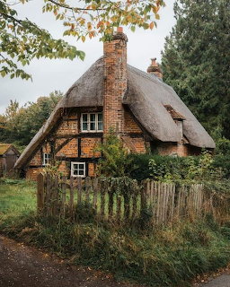 Rustic brick and timber cottage with a thatched roof behind a wooden fence