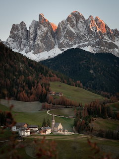 Alpine village with church below jagged Dolomite mountain peaks at sunset with autumn foliage