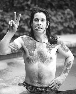Black and white photo of shirtless tatooed man with long dark hair in a pool making a peace sign