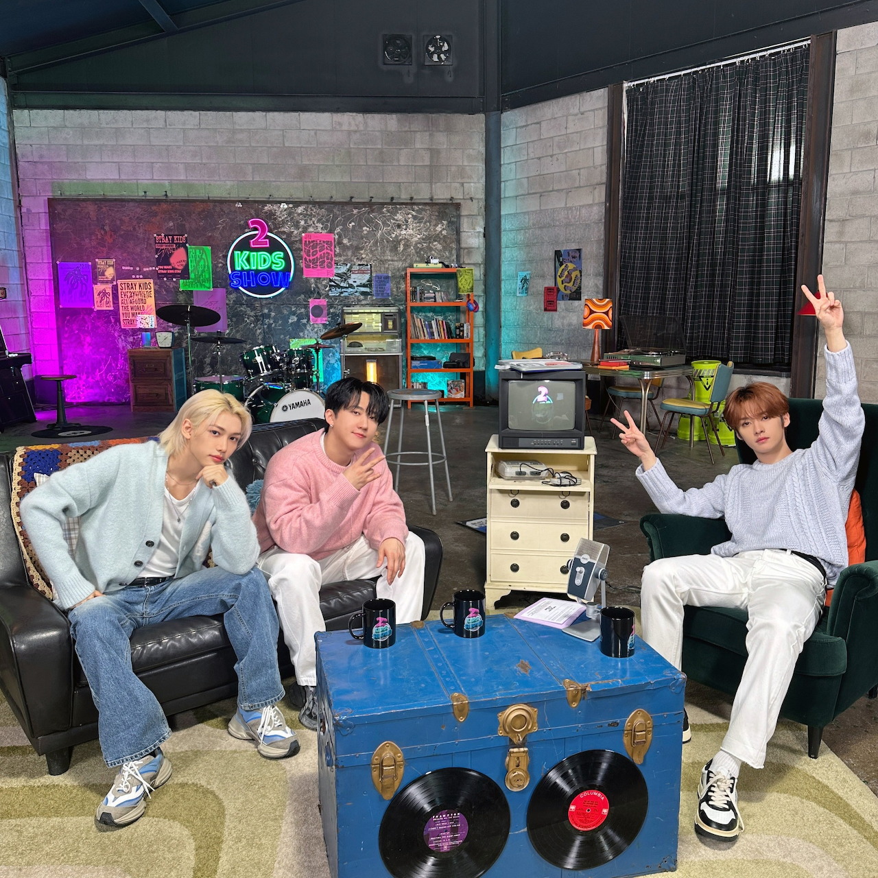 Three young men posing in a colorful studio room with chairs, records, and a vintage TV