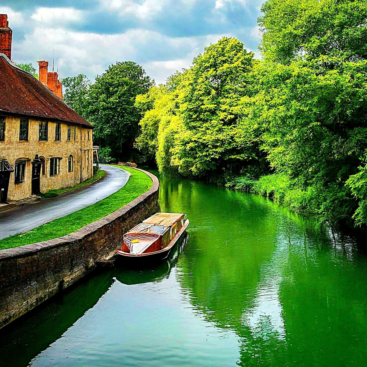 Wooden rowboat on peaceful green canal beside yellow stone cottages with trees reflecting in water