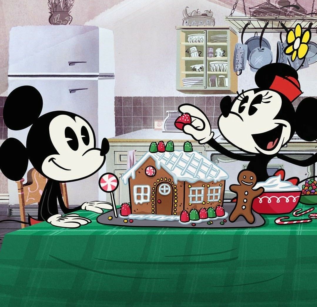 Mickey and Minnie Mouse decorating a gingerbread house on a green table in a kitchen