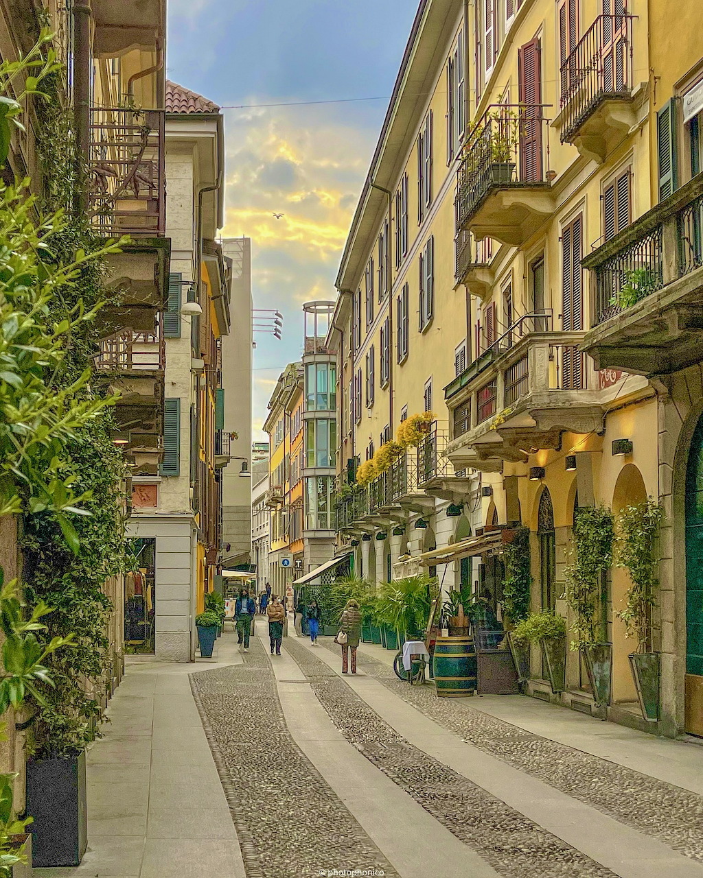 Narrow European street with colorful buildings, balconies, shops, and patterned cobblestone pavement at dusk