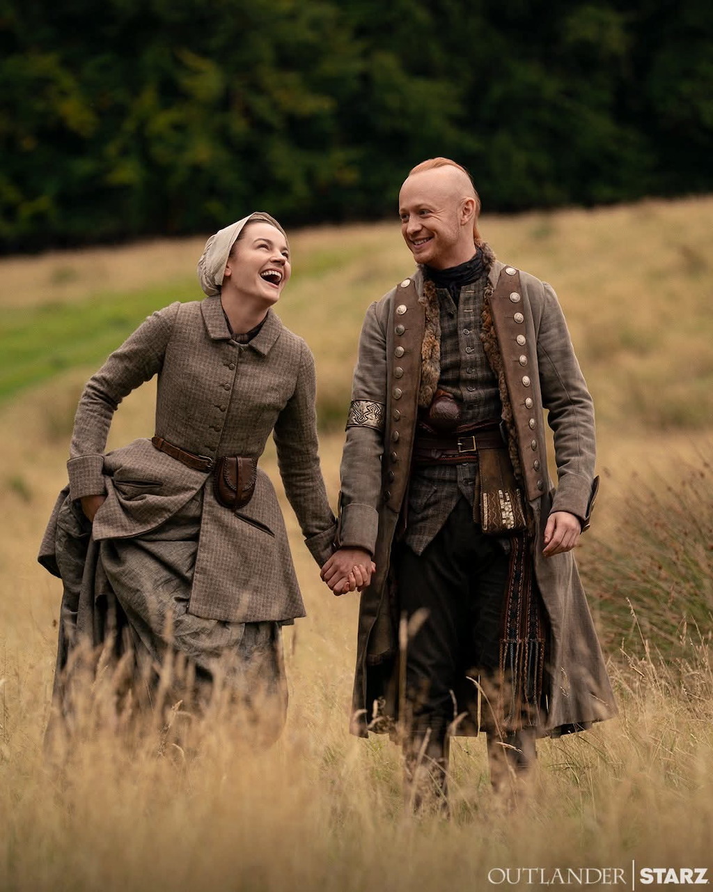 Man and woman in period costumes holding hands and laughing while walking through a grassy field