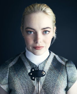 Portrait of Emma Stone with blonde hair, blue eyes, wearing a plaid jacket and white turtleneck