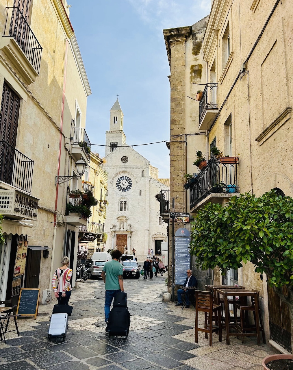 Sunny Italian street with white church, tall buildings, potted plants, and cafe tables