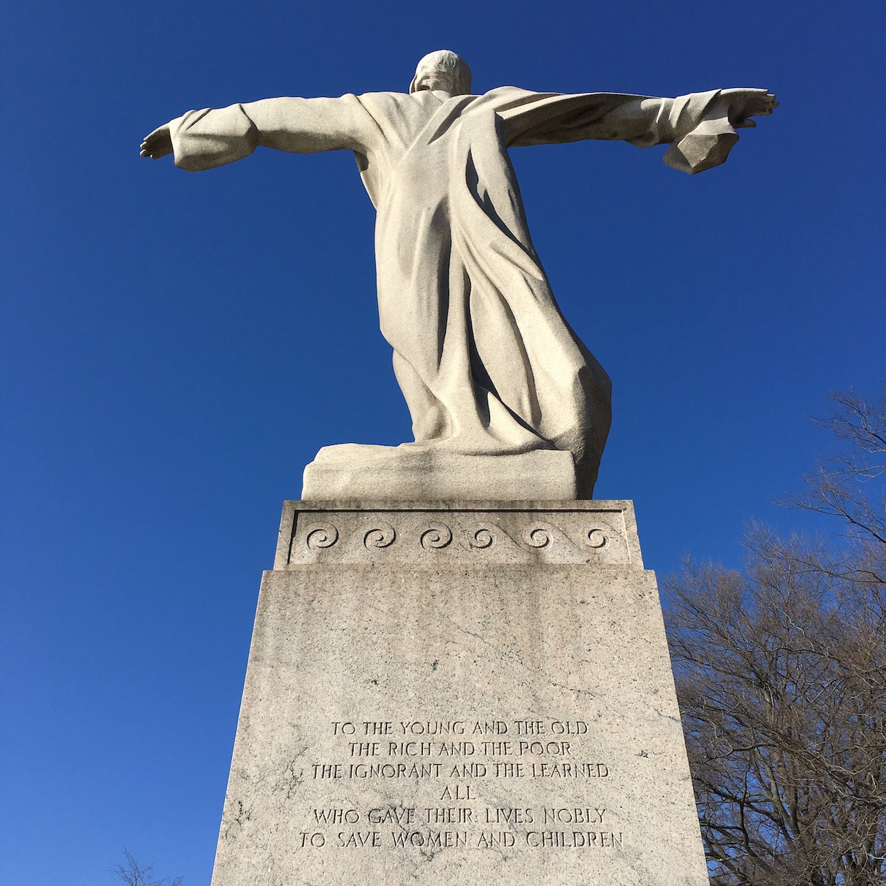 White marble memorial statue with outstretched arms on stone pedestal against blue sky