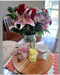 Glass vase with pink lilies, red roses and purple flowers on a wooden tray with cards and a crucifix on a table