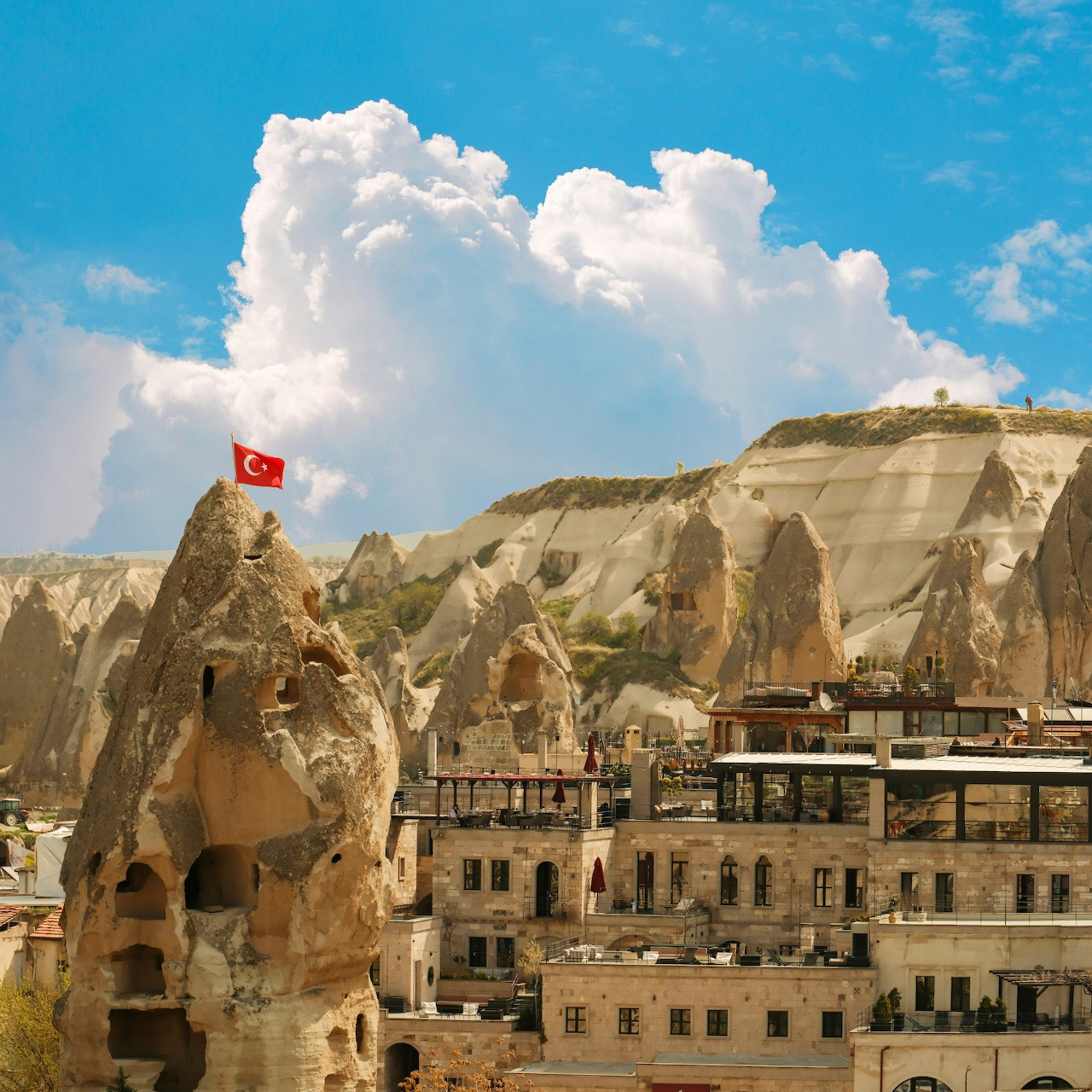 Historic cave dwellings carved into beige rock formations in Cappadocia with Turkish flag and blue sky