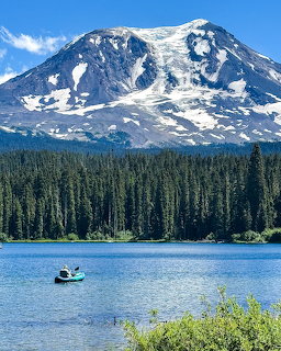Snow-covered mountain peak above evergreen forest and blue lake with kayakers under clear sky