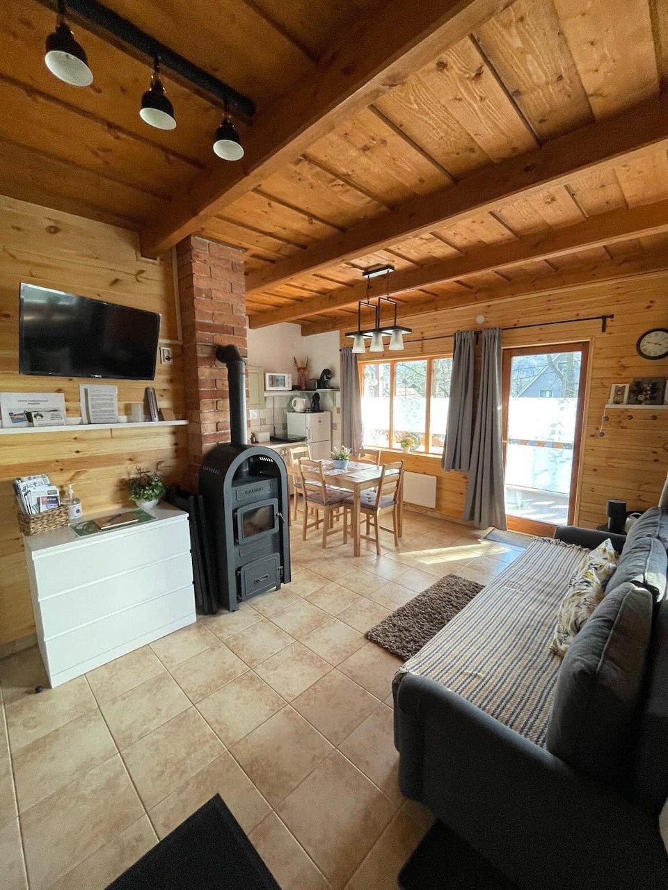 Cozy log cabin living room with wood ceiling, black stove, gray sofa, and dining table near a window