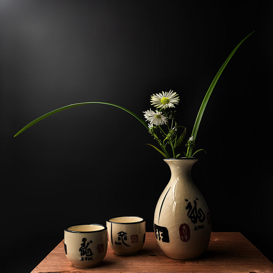 Small white flowers in a ceramic vase with two cups on a wooden table against a black background