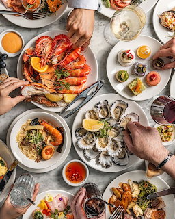 Overhead flatlay of seafood brunch with lobster, oysters, salmon, pastries and hands reaching for food on white table