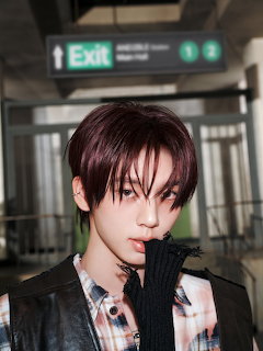 Young man with dark red hair, plaid shirt, and black glove posing indoors under an exit sign.