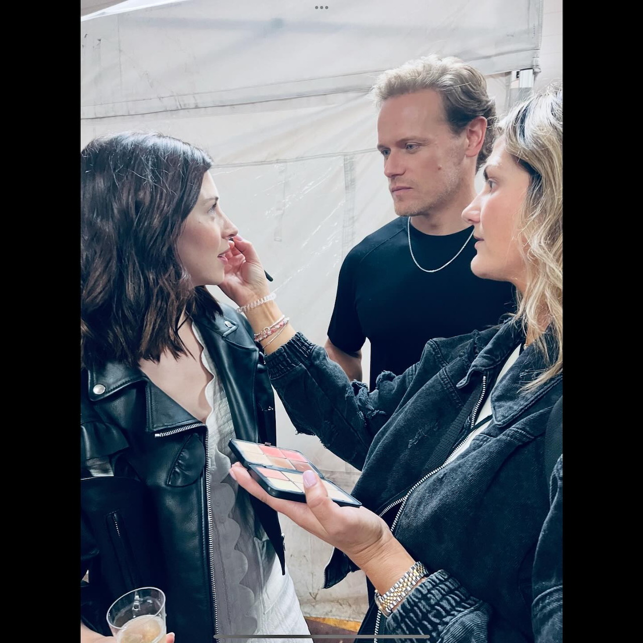 Three people talking indoors, with one woman touching another woman's face and holding a phone