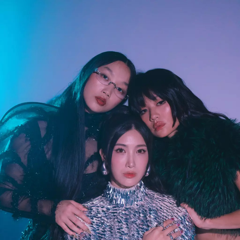 Three women in dark fashion outfits posed together with blue-purple lighting