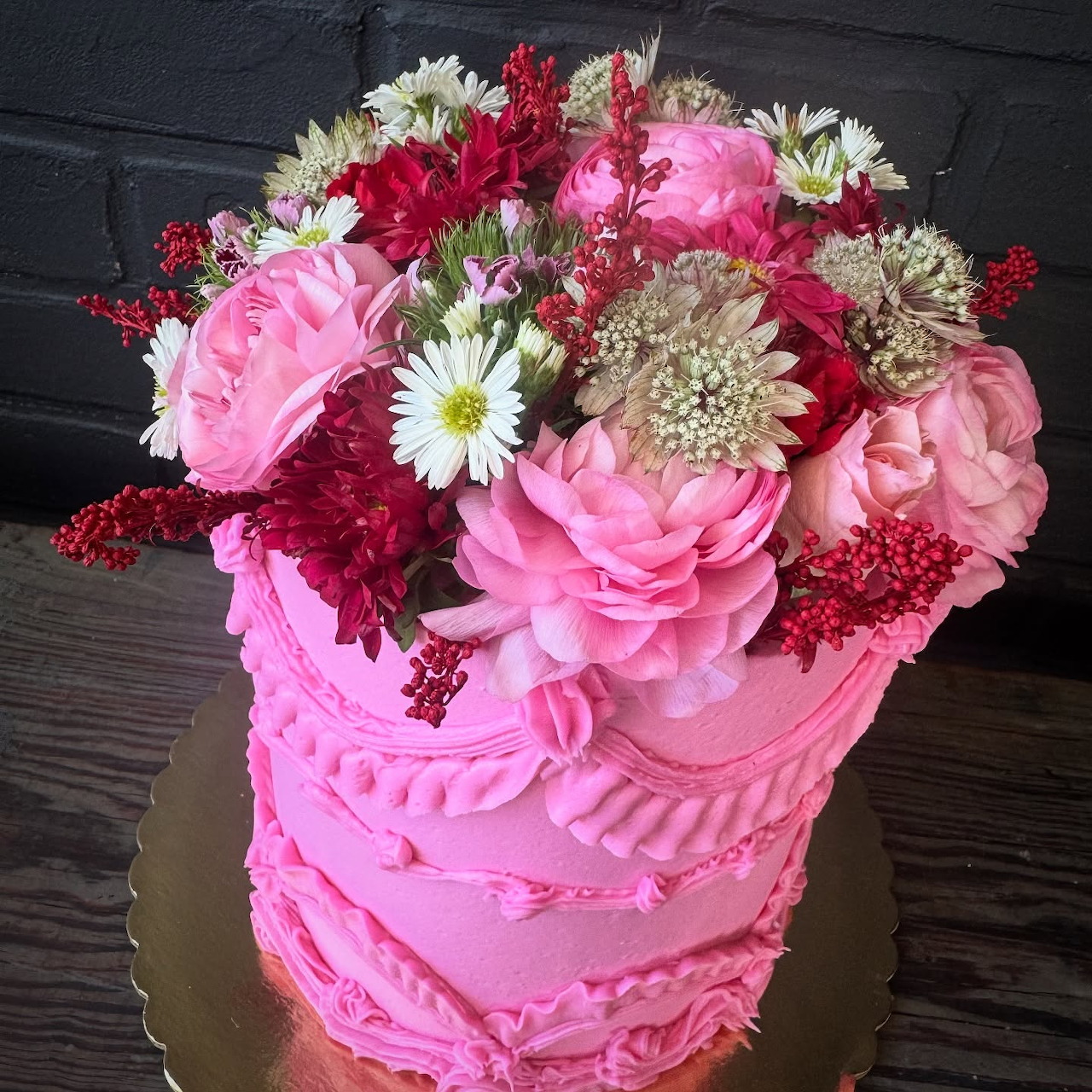 Pink frosted cake decorated with mixed flowers on a gold cake board