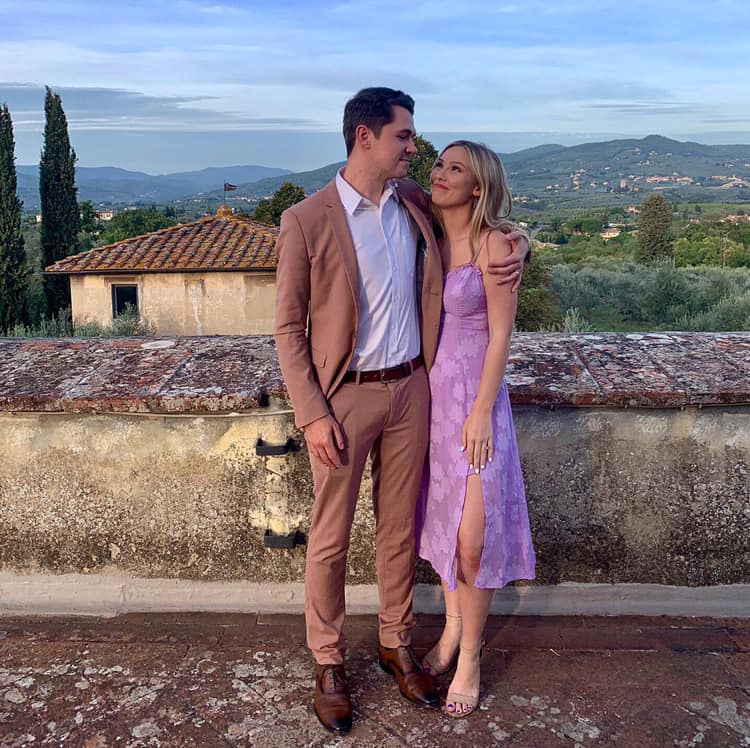 A dressed-up couple standing together on a terrace with hills and a rustic building behind them.