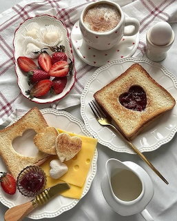 Breakfast table with heart-shaped toast, jam, strawberries, cheese, cappuccino, and white dishes on plaid cloth